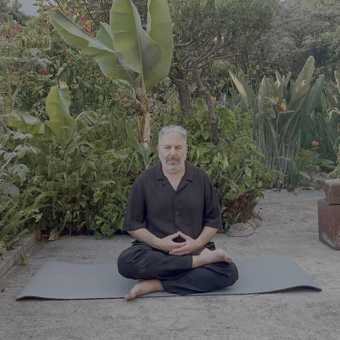 Robert sitting cross-legged on a mat in a garden of ferns and banana leaves