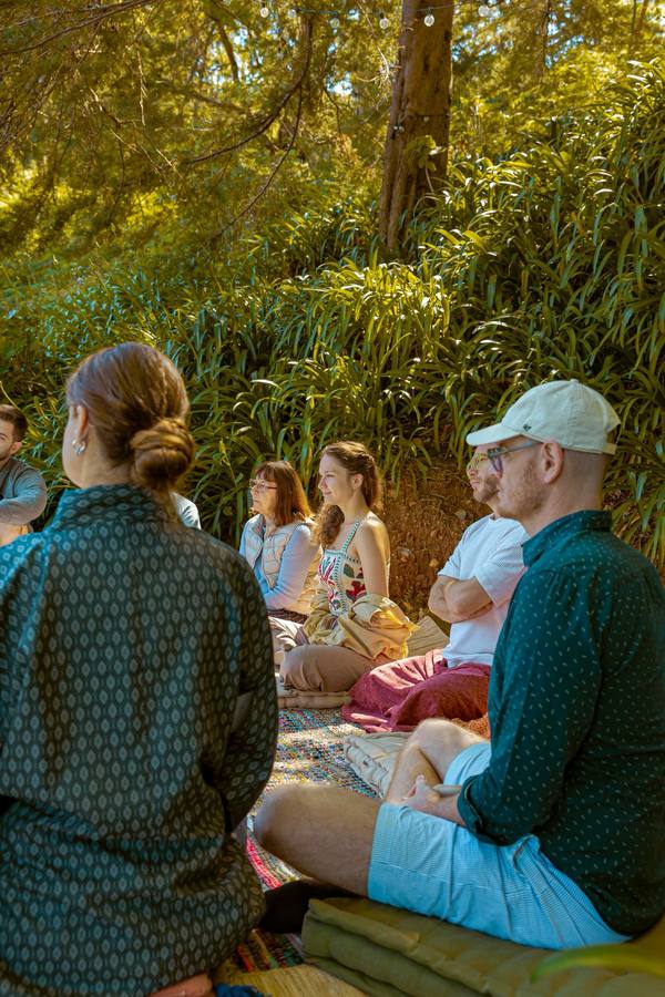 A workshop circle sitting on cushions and rugs under trees, in dappled afternoon light